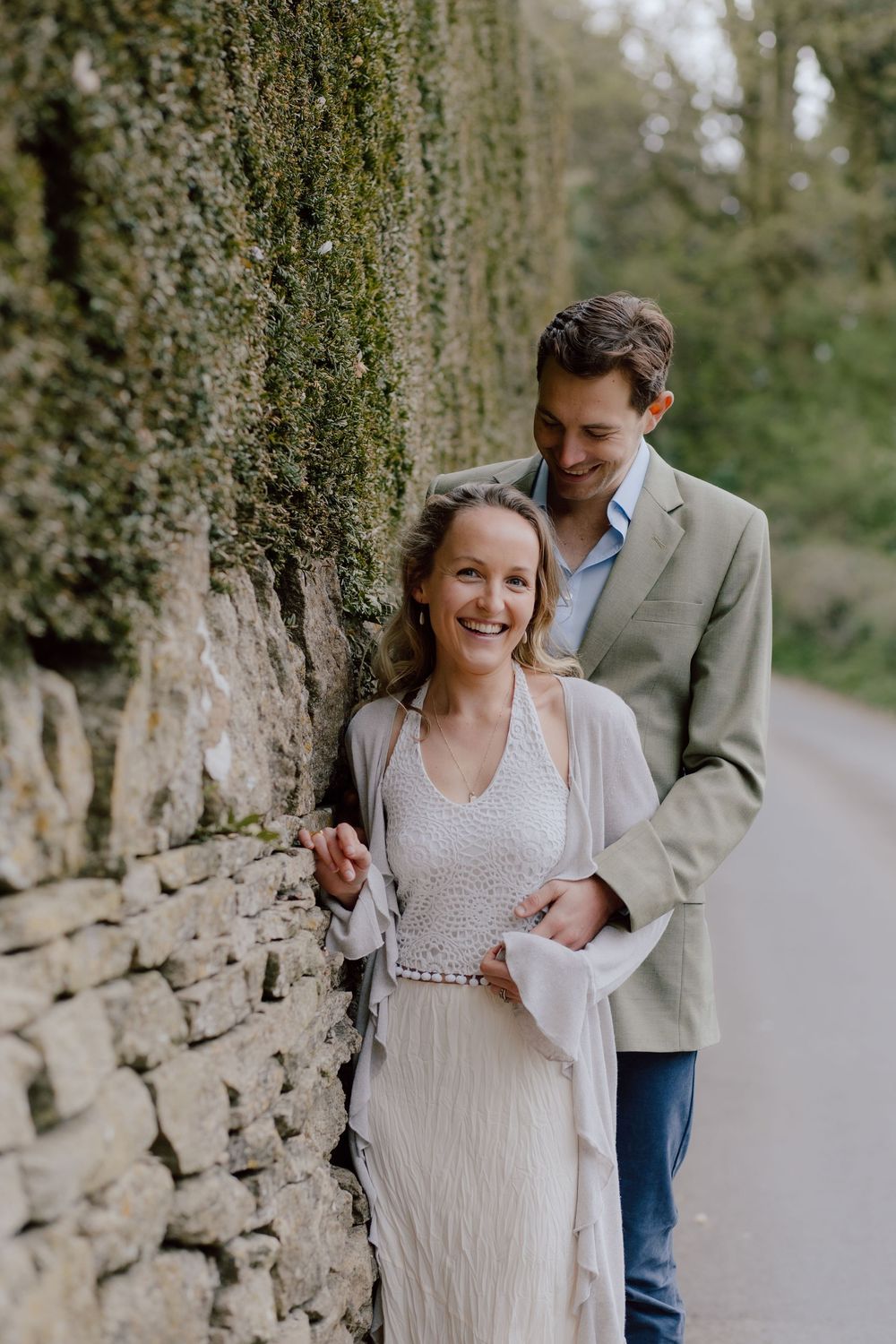 A loving couple shares an intimate moment against a stone wall with overhanging moss and greenery on a garden path.