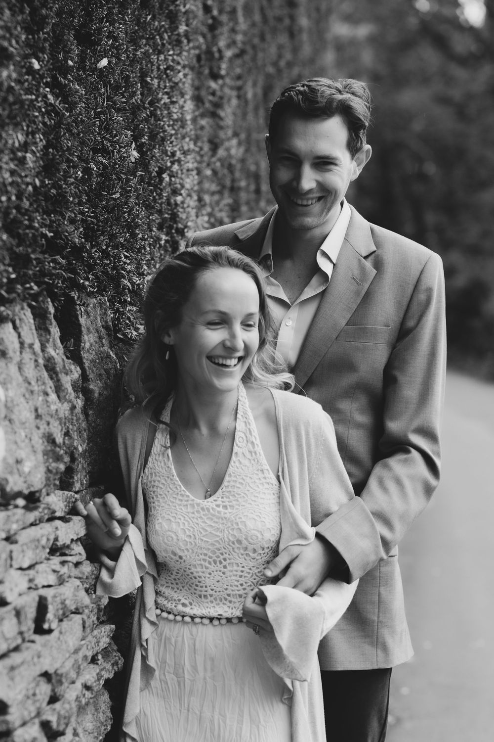 A happy couple shares a joyful moment together against a stone wall in this black and white romantic portrait.
