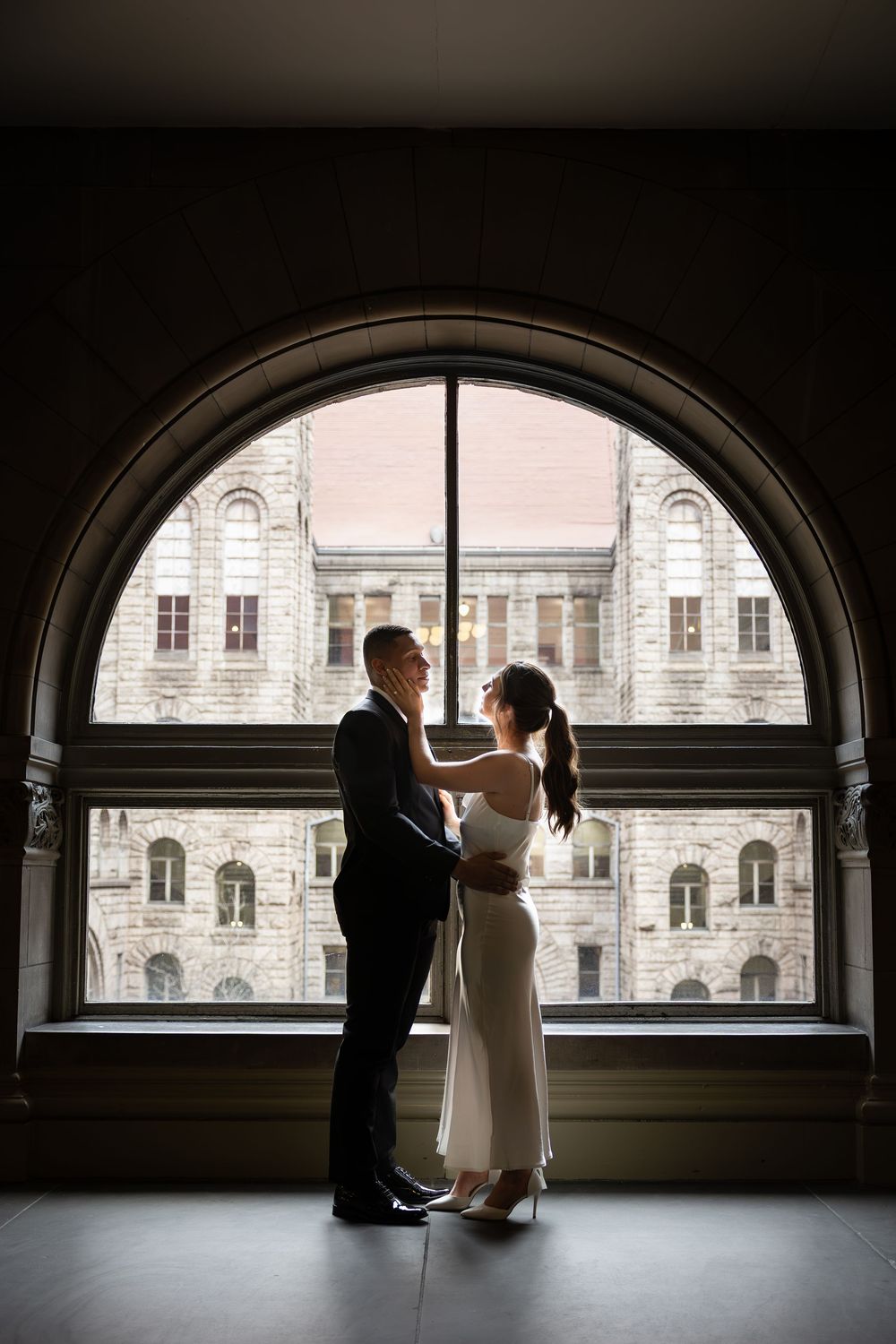 Silhouetted couple embracing in front of large arched window with historic architecture view.