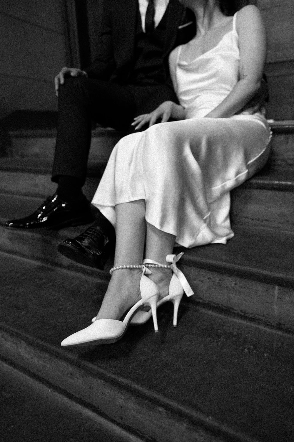 Black and white photo showing elegant formal attire with white heels and black dress shoes on stone steps.