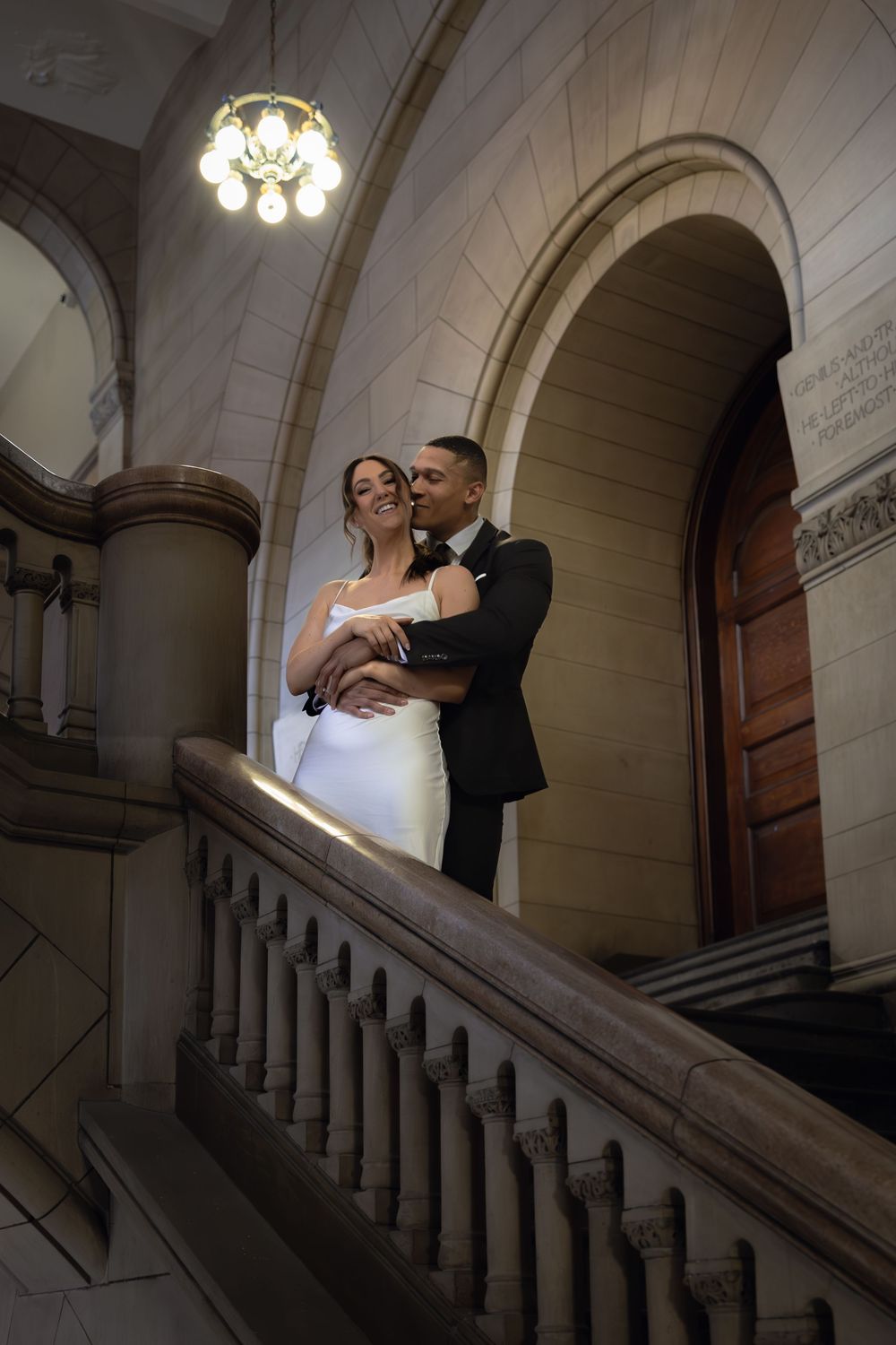 A couple in wedding attire stands intimately on an elegant marble staircase inside a historic building with arched architecture.