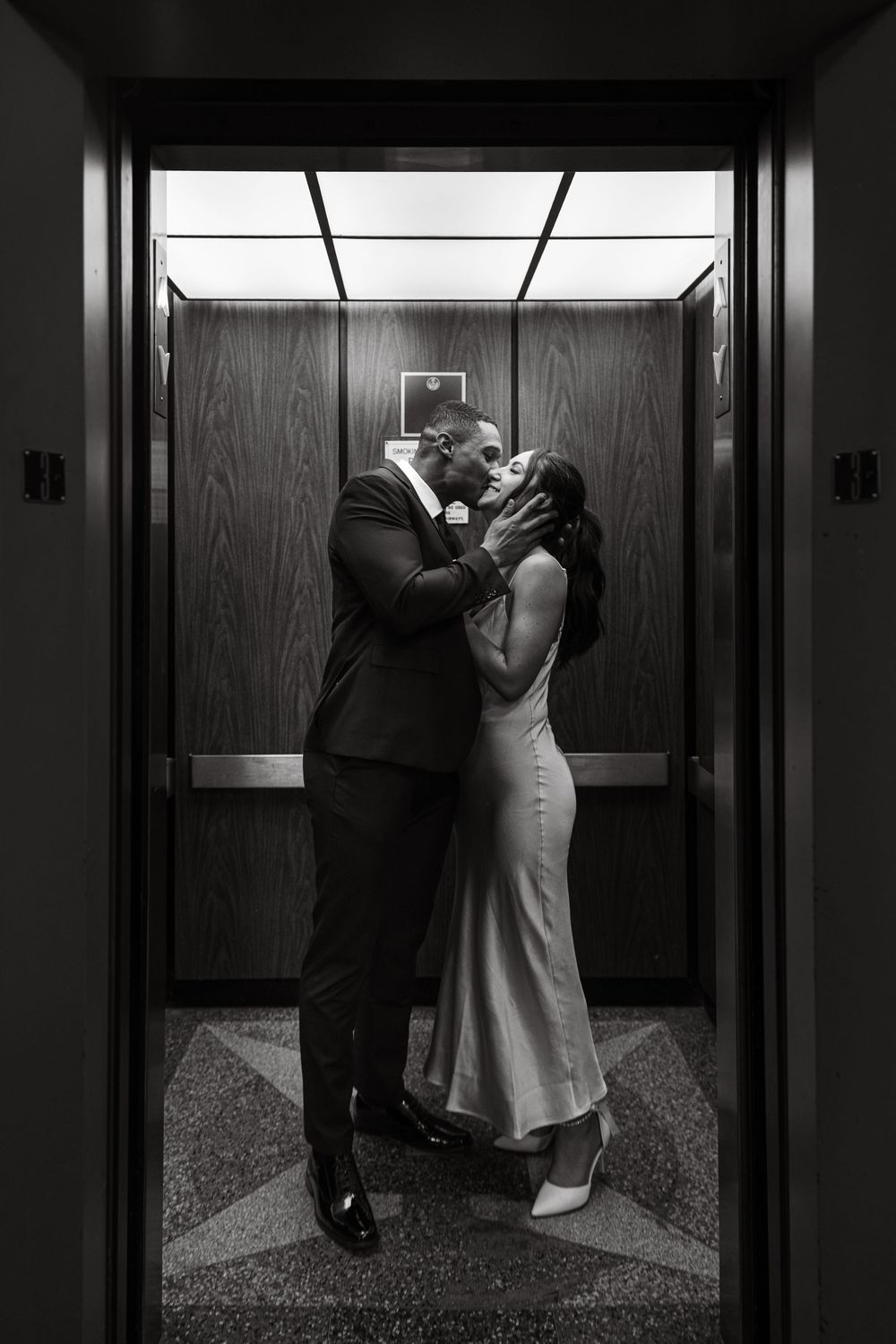 A romantic black and white photo of a couple sharing a kiss in an elevator with wood paneled walls.