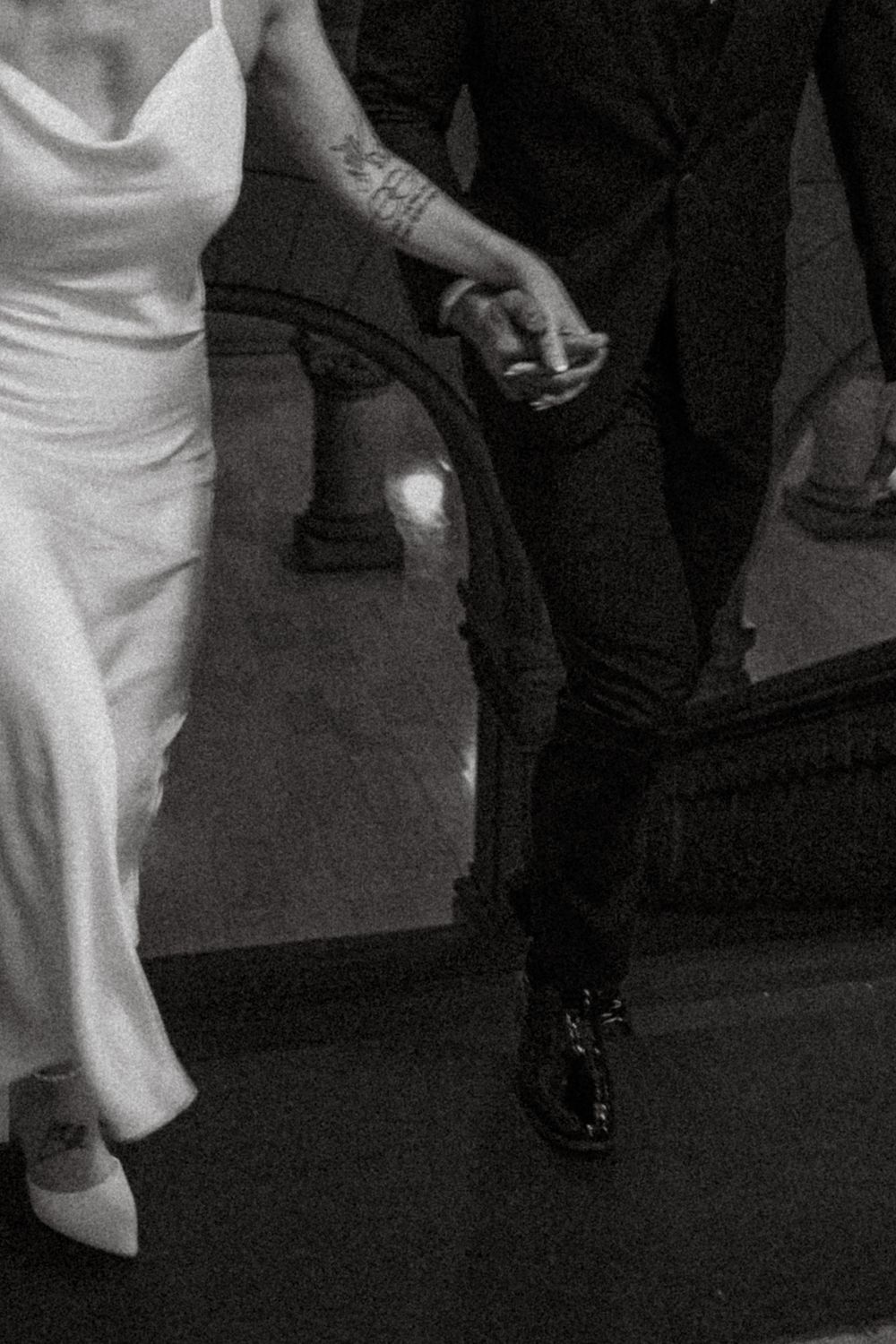 Two people holding hands at a formal event, with bride wearing white dress and groom in dark suit and dress shoes.