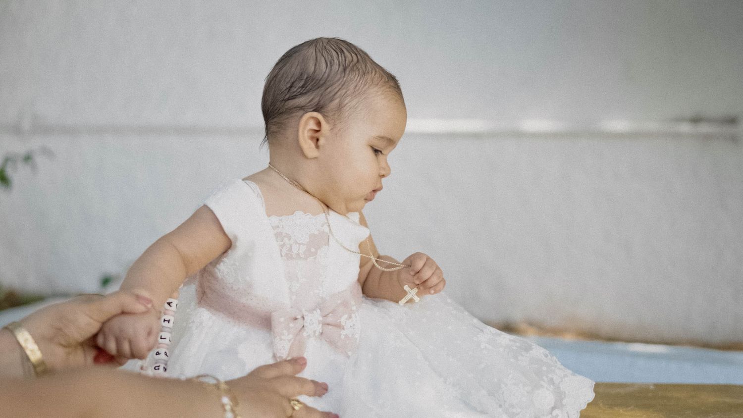 A small baby wearing an elegant white lacy christening gown sits on a light colored sofa.