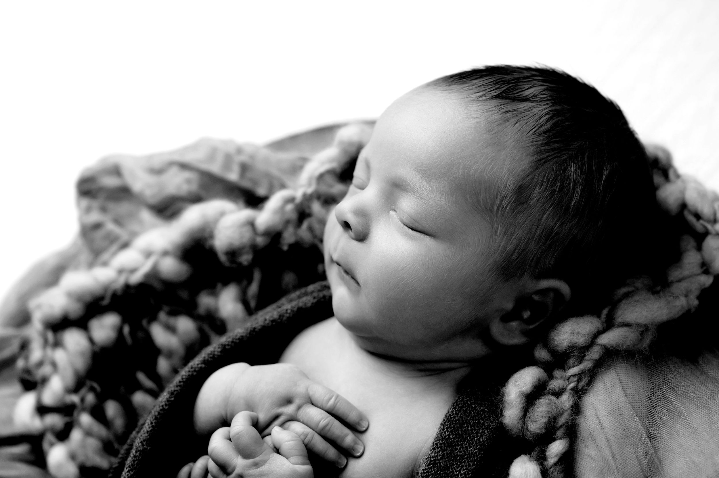 Sleeping newborn baby wrapped in soft white swaddle blanket resting on cream textured backdrop during photography session.