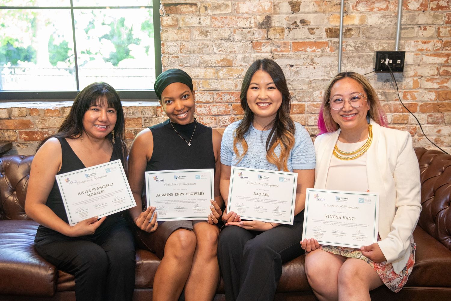 A group of students smiling while sitting on a brown leather couch and displaying their achievement certificates.