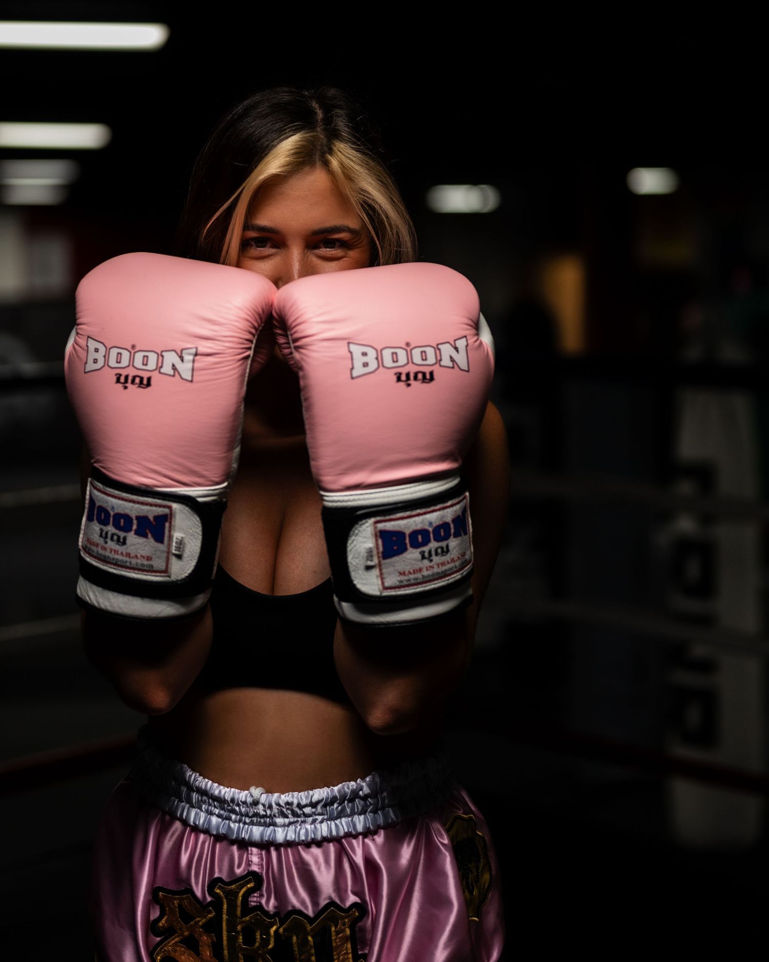 Boxer in black sports bra and pink shorts holds up pink boxing gloves in dark gym setting.