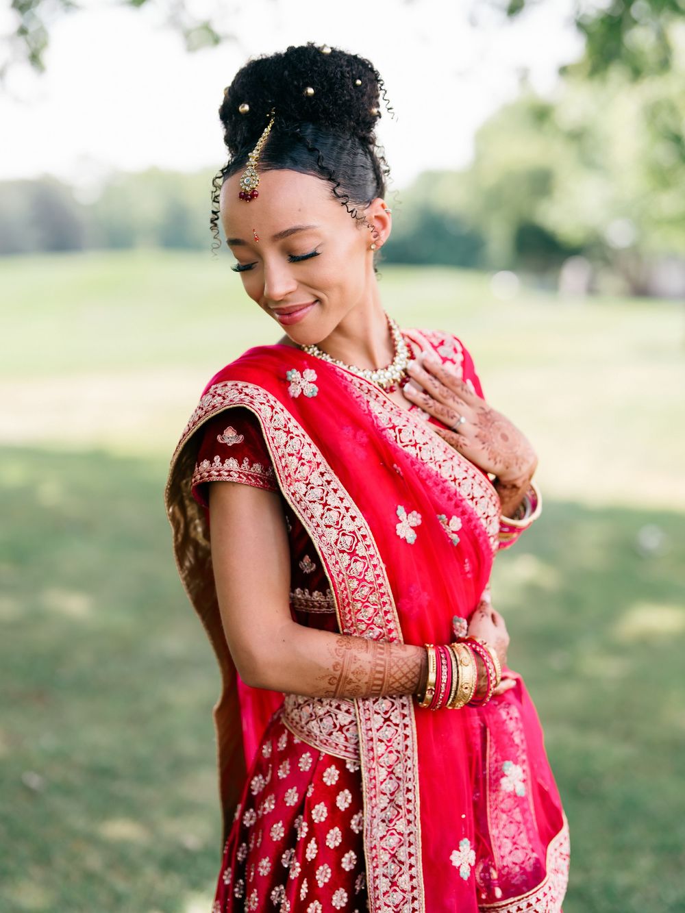 A person wearing an elegant red and gold embroidered sari and traditional Indian bridal jewelry smiles joyfully.