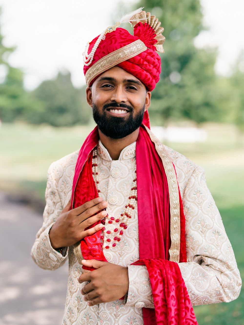 Traditional Indian wedding attire features an ornate cream sherwani with red turban and scarf in an outdoor setting.