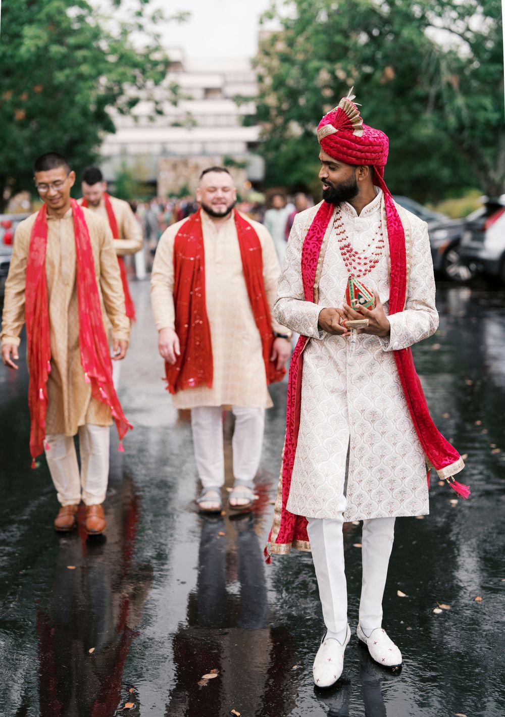 Three grooms in traditional Indian wedding attire walking on wet pavement wearing red and white ceremonial outfits.