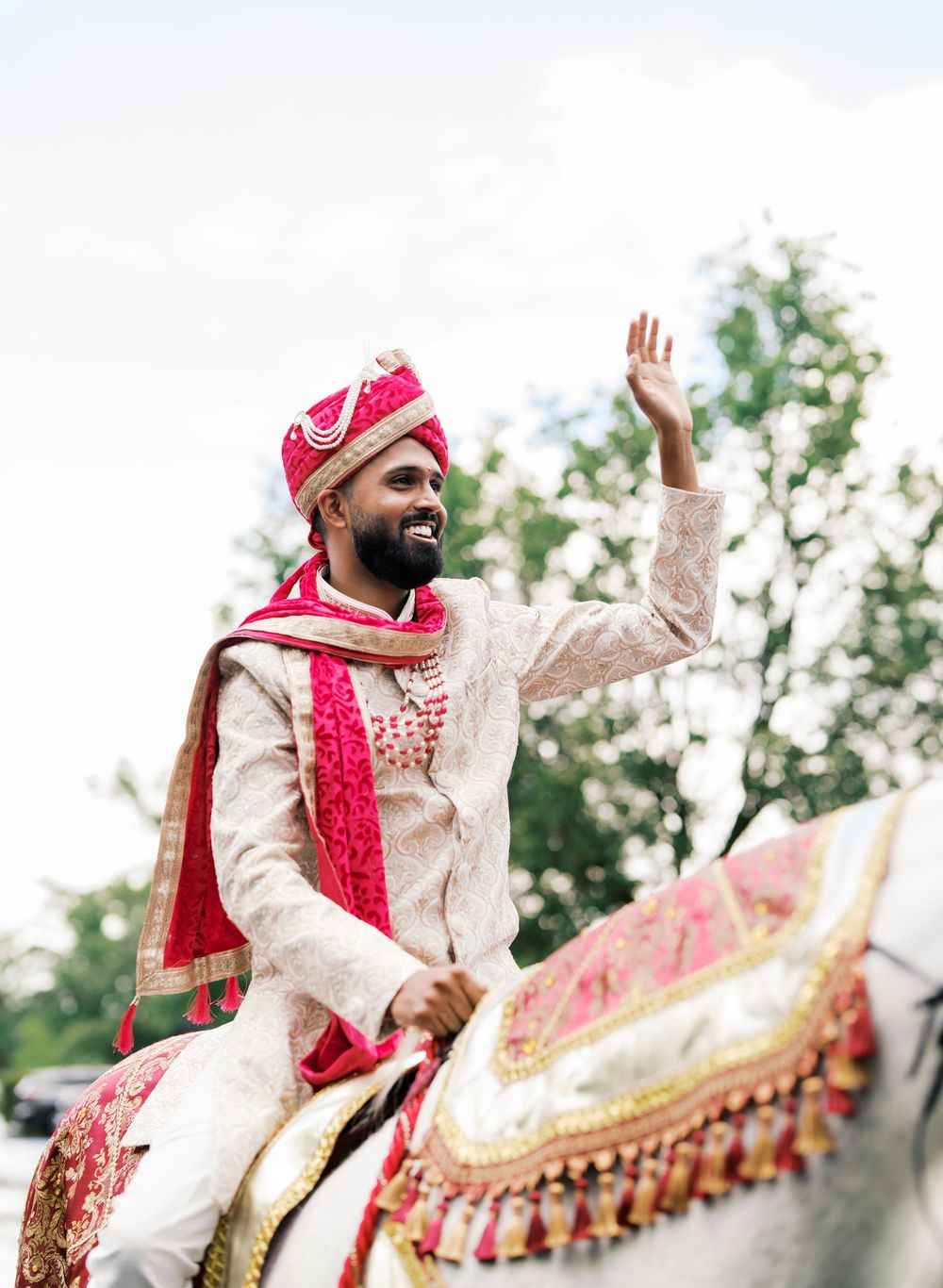 A traditionally dressed groom in cream and red attire waves while seated on decorated horse.