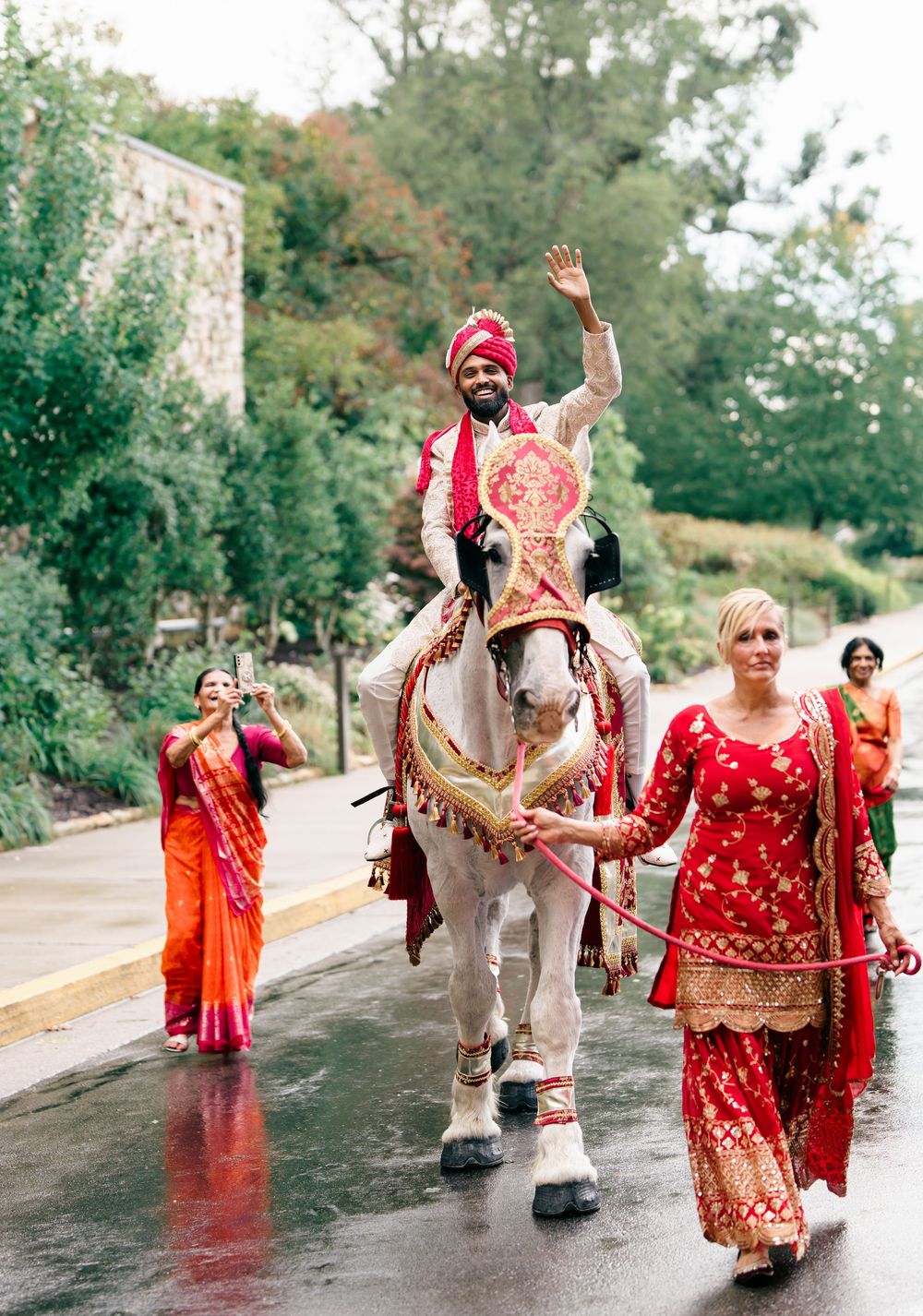 Traditional Indian baraat wedding procession with decorated white horse and guests in colorful attire on rainy street.