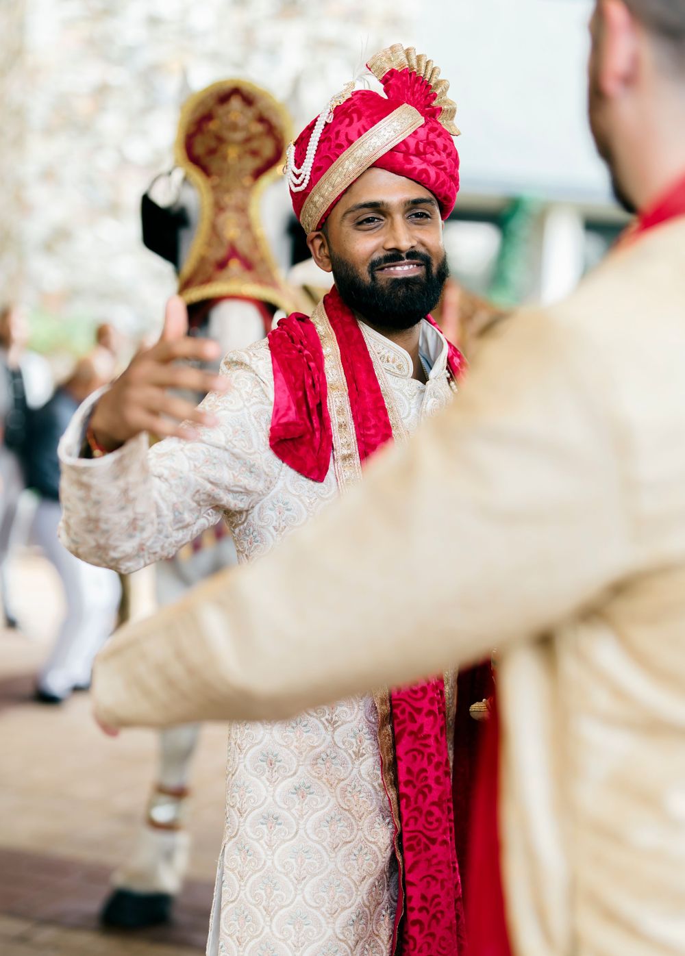 Indian groom in ornate white and red traditional wedding attire smiling during celebration.