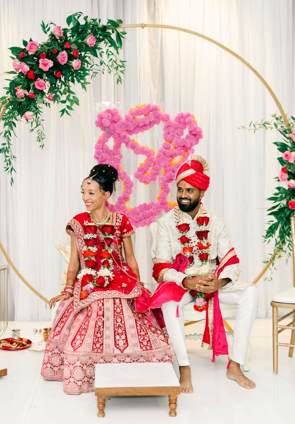Couple in traditional wedding attire seated at ceremony with gold hoop arch and pink floral decorations.