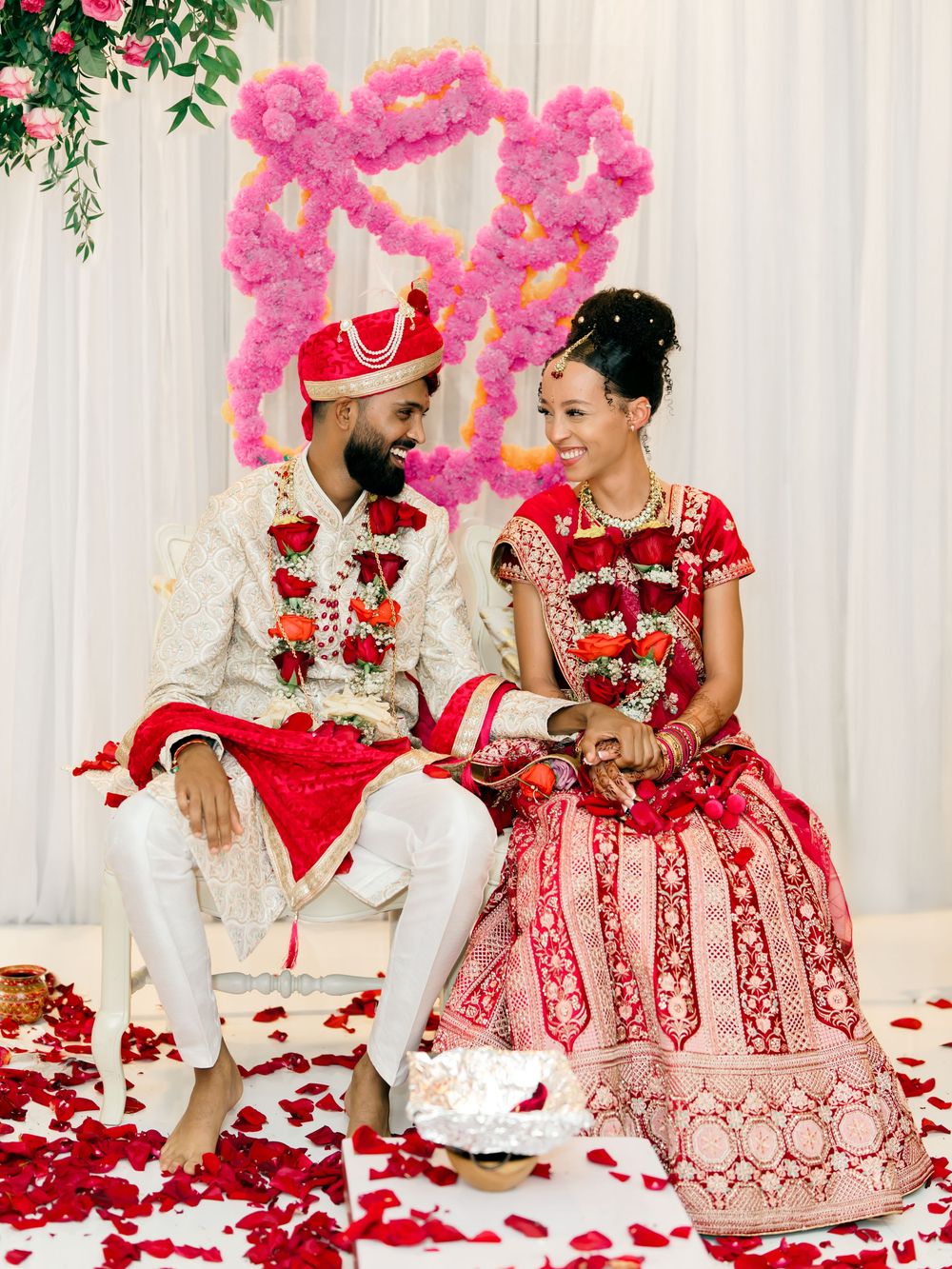 Traditional Indian wedding ceremony with couple in red and white outfits seated under pink floral heart decoration.