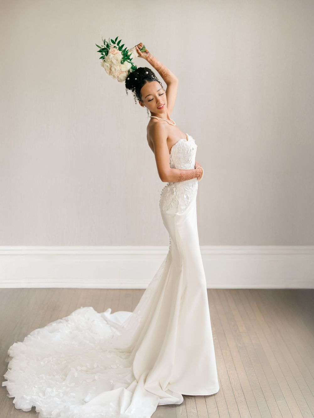 Elegant bride in form-fitting white lace gown with sweeping train poses gracefully with raised arm against neutral backdrop.