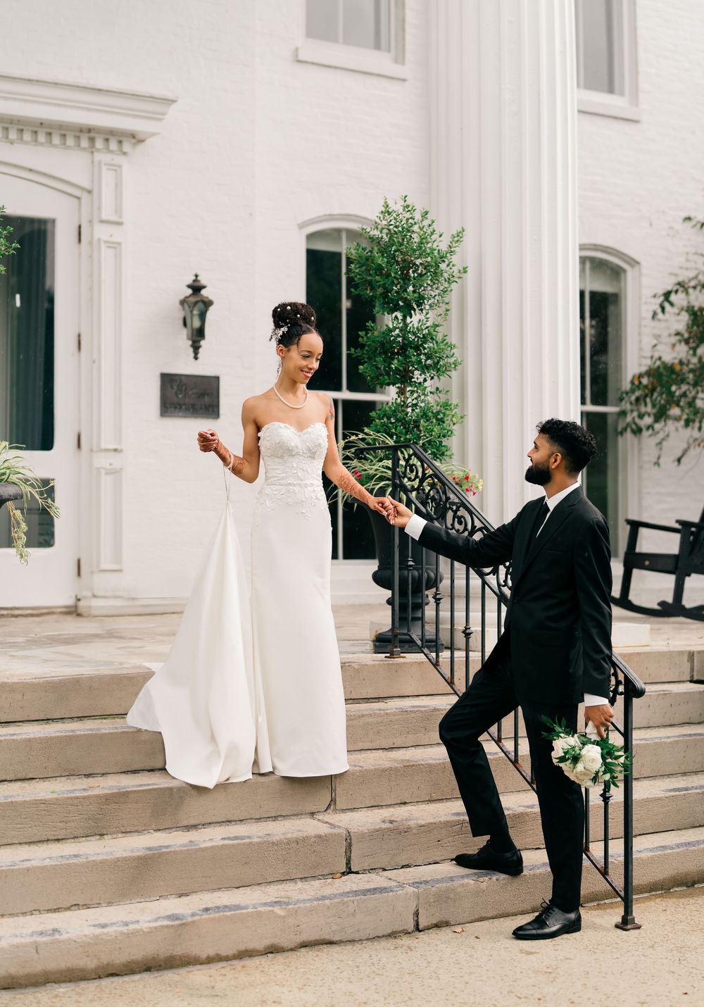 A couple in wedding attire poses on white stone steps, with the groom offering his hand to the bride.