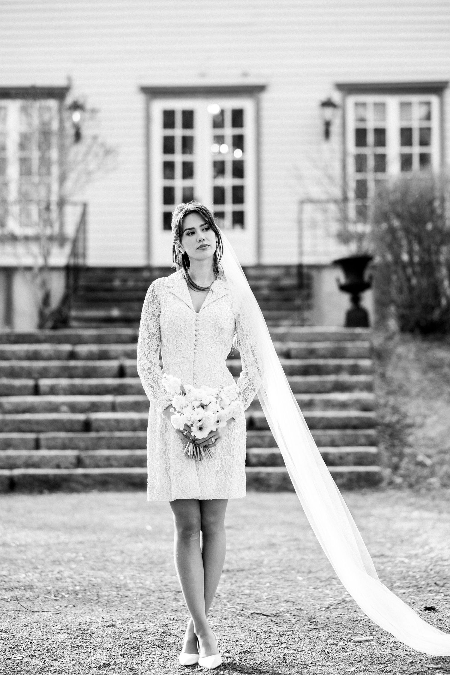A bride in a short lace dress with flowing veil stands on stone steps outside an elegant white mansion.