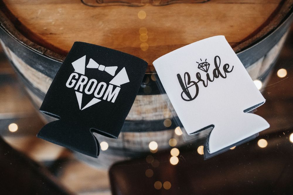 Black and white bride and groom wedding koozie drink holders displayed on wooden barrel with bokeh lights in background.