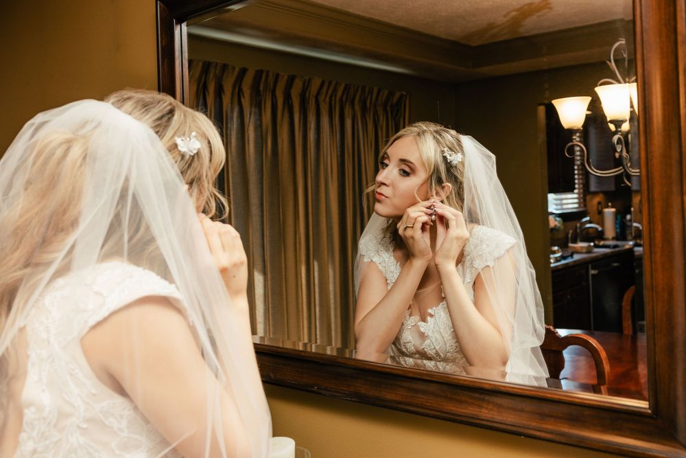 Person in bridal attire puts on earrings while looking in an ornate mirror.