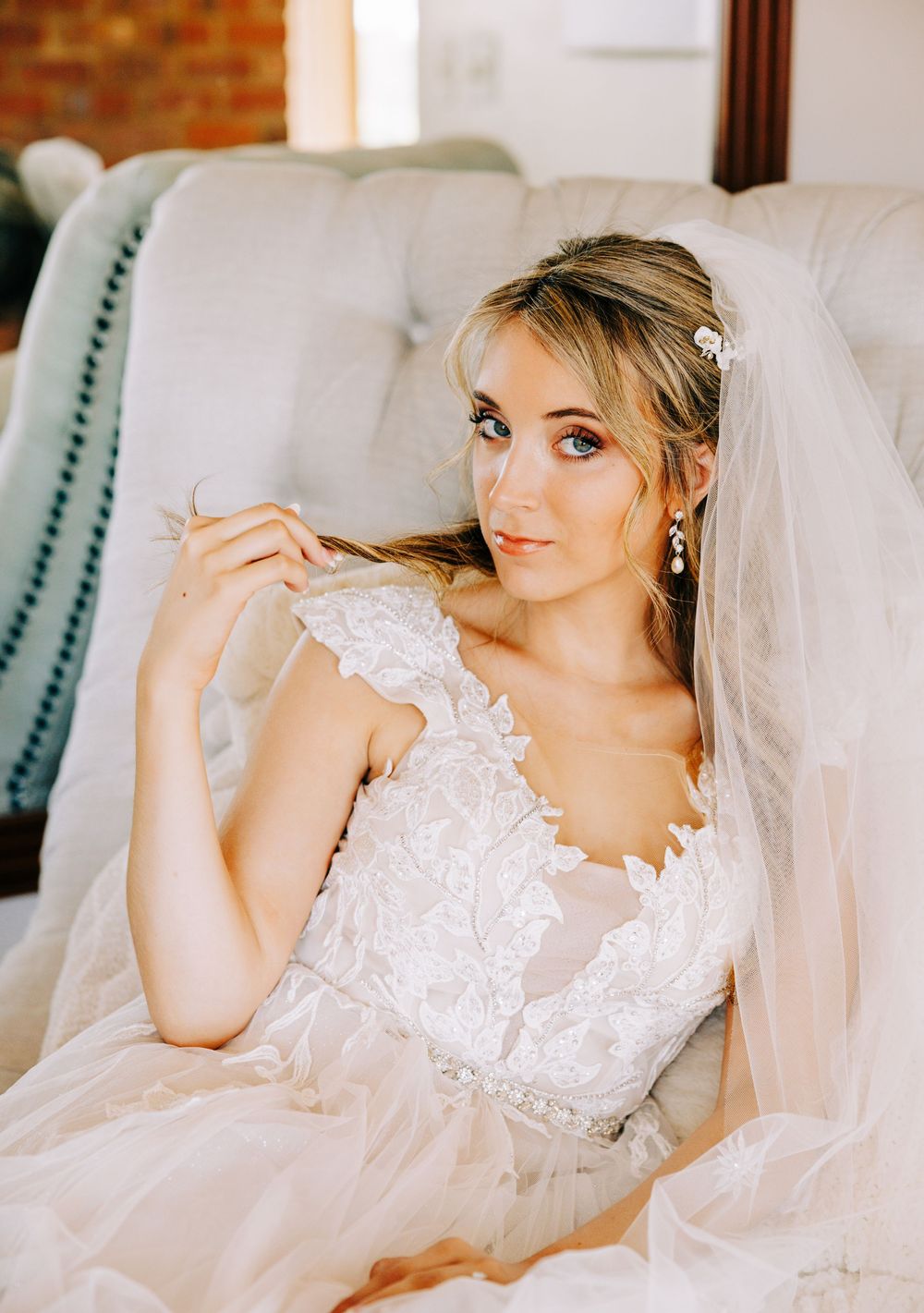 A bride in a delicate lace wedding dress with veil reclines on a white sofa against a brick wall backdrop.