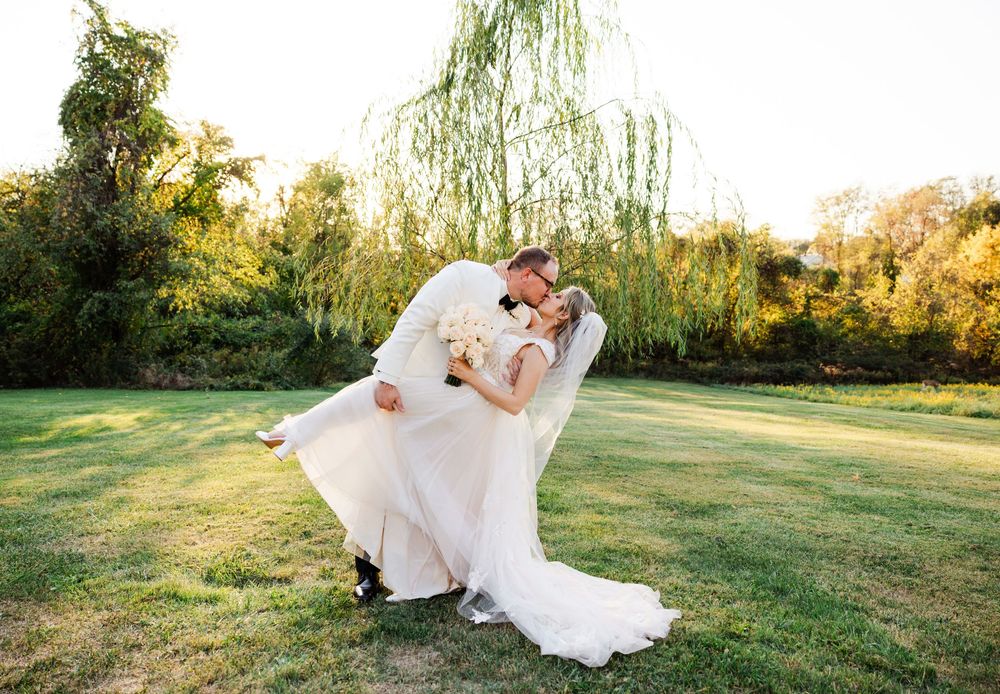 A romantic couple embraces on a sunlit lawn with a flowing white wedding dress and weeping willow tree in the background.
