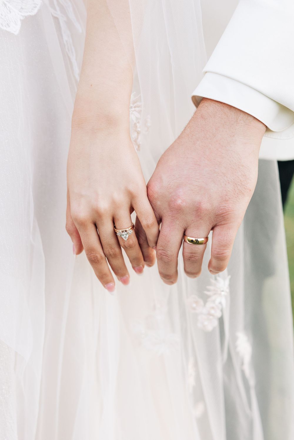 Close-up of newlywed couple's hands with wedding rings against white bridal dress and veil.