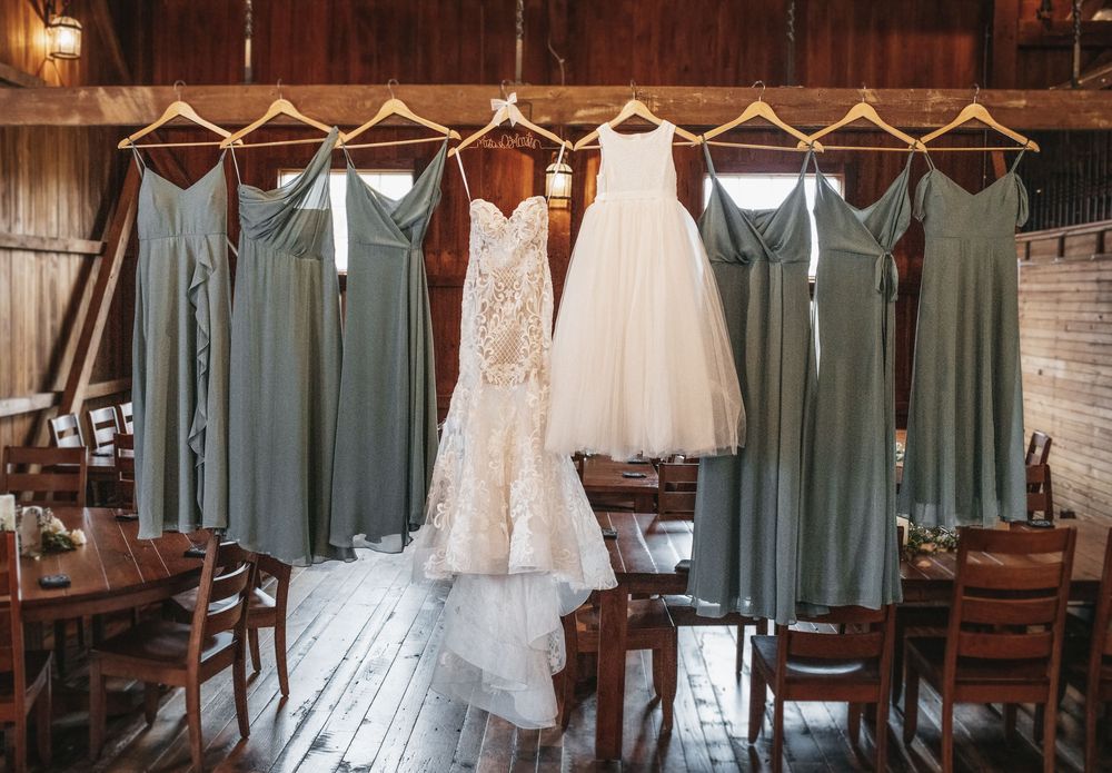 Elegant wedding gown and sage green bridesmaid dresses hanging in a rustic wooden barn setting.