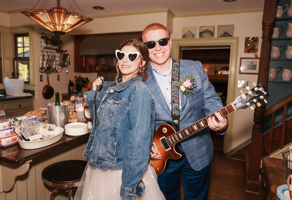 Happy couple wearing sunglasses pose at a restaurant, with one holding a ukulele and wearing a denim jacket and blazer.
