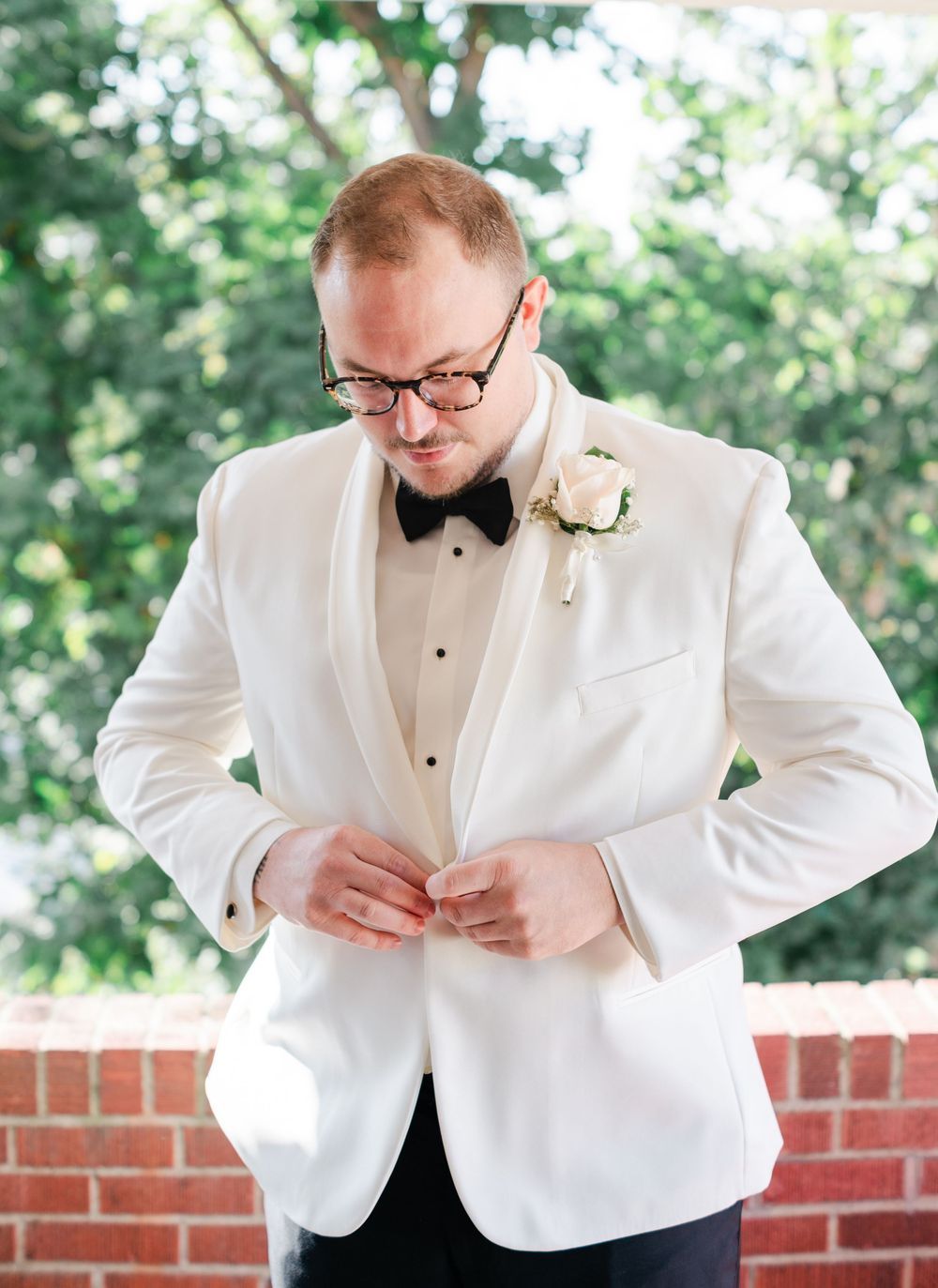 Person in white tuxedo jacket and bow tie adjusts formal attire against brick wall backdrop.