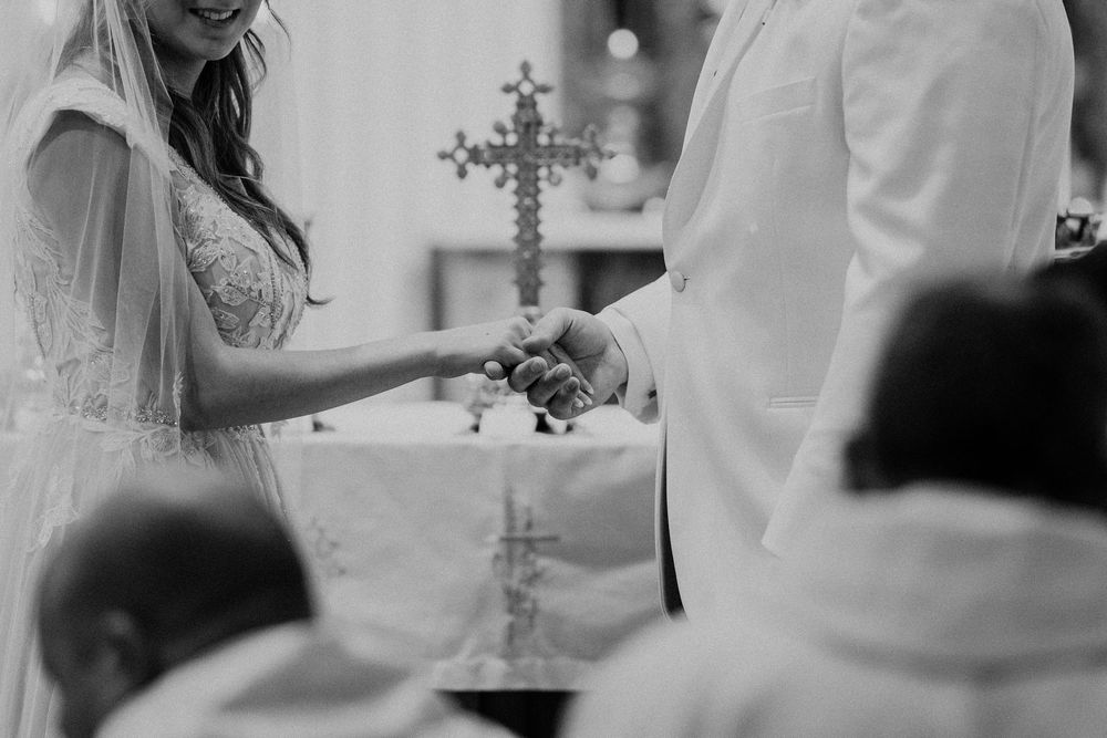 A black and white wedding ceremony photo showing exchanging of rings with an ornate cross in the background.