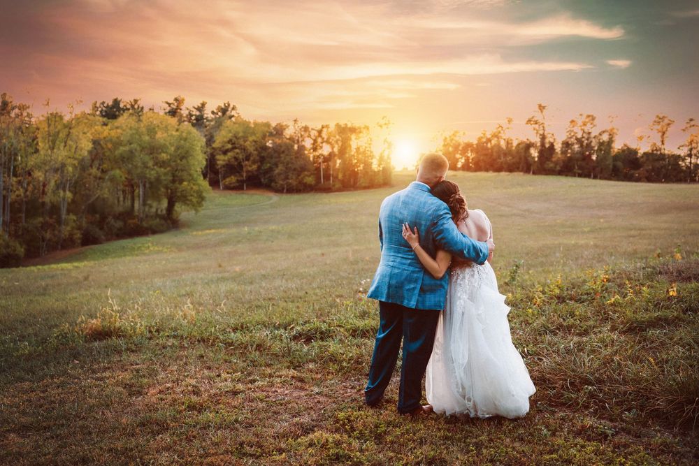 A couple embraces while watching a golden sunset over a serene meadow with trees in the background.