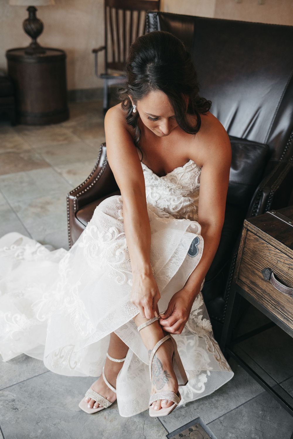 Bride in a white lace wedding dress fastens delicate strappy sandals while sitting in a leather chair.