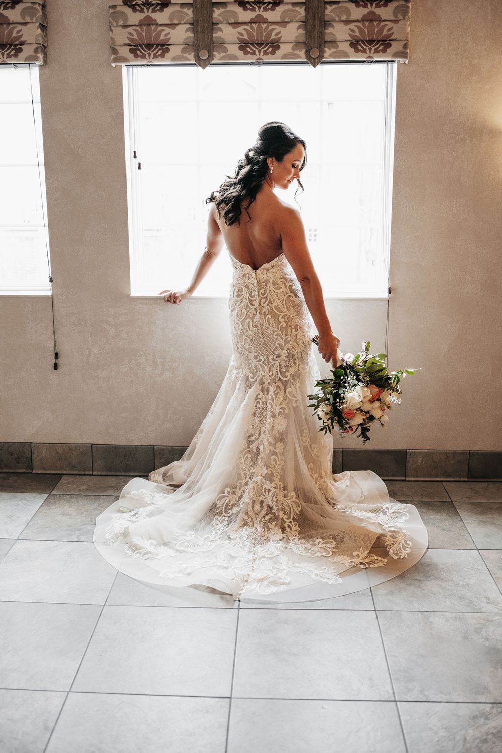 An elegant bride in a backless lace wedding gown with a flowing train poses near a window while holding a floral bouquet.