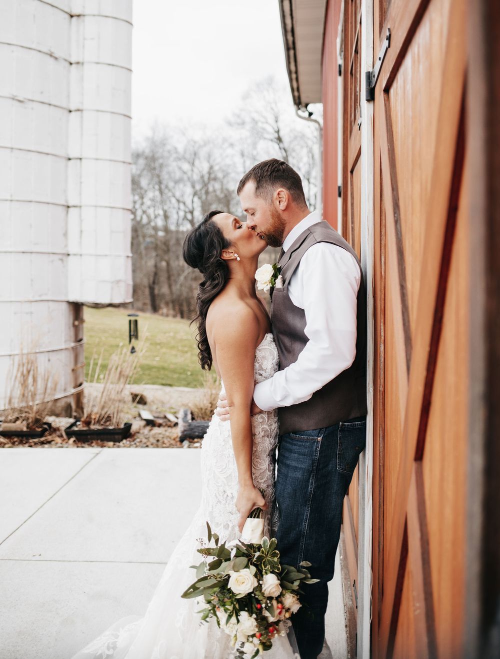 A couple shares a romantic kiss against a rustic barn wall while holding a white floral bouquet.