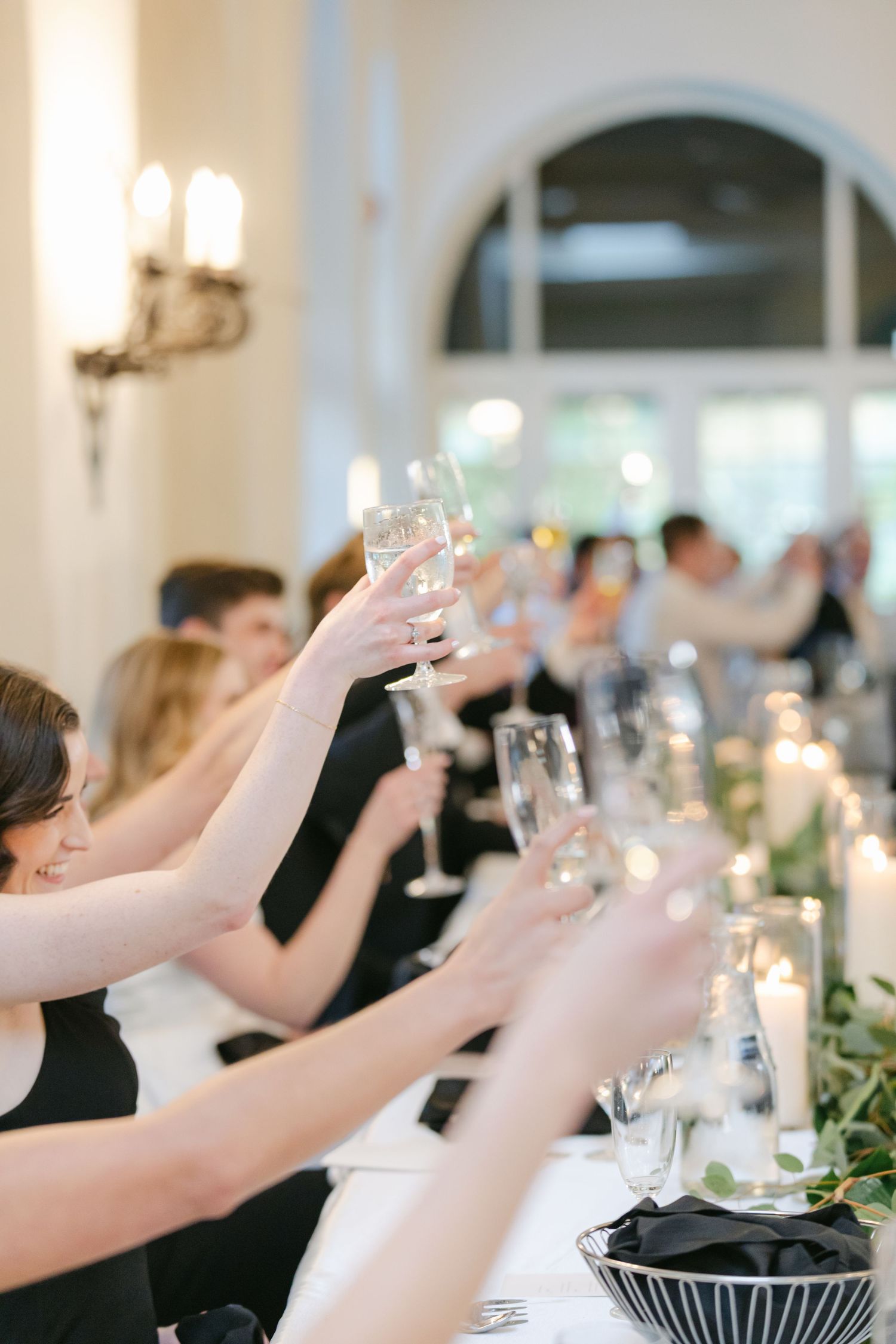 Wedding guests raise their glasses in a toast during an elegant reception in a room with arched windows and candlelight.