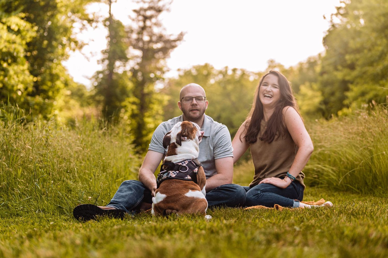 A couple sits in a sunlit meadow with their Boston Terrier dog during a warm summer evening.