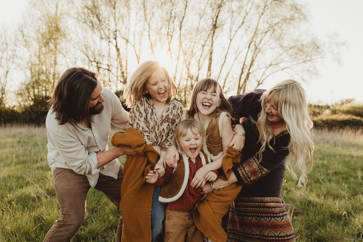 A family shares a playful moment together in a sunlit field with tall grass during golden hour.