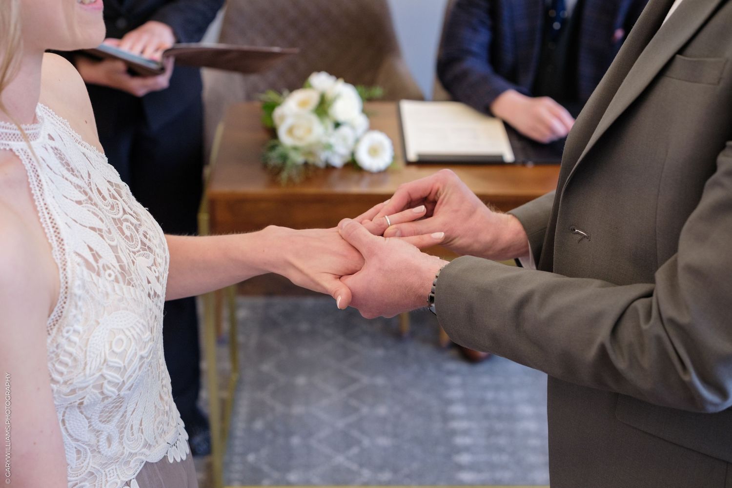 Close-up of hands exchanging wedding rings during a ceremony with white floral bouquet in the background.