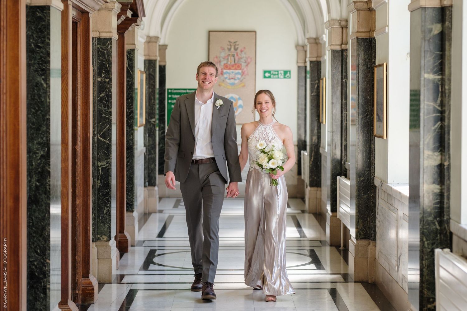 A couple walks together in formal attire through an elegant hallway with marble columns and floors.