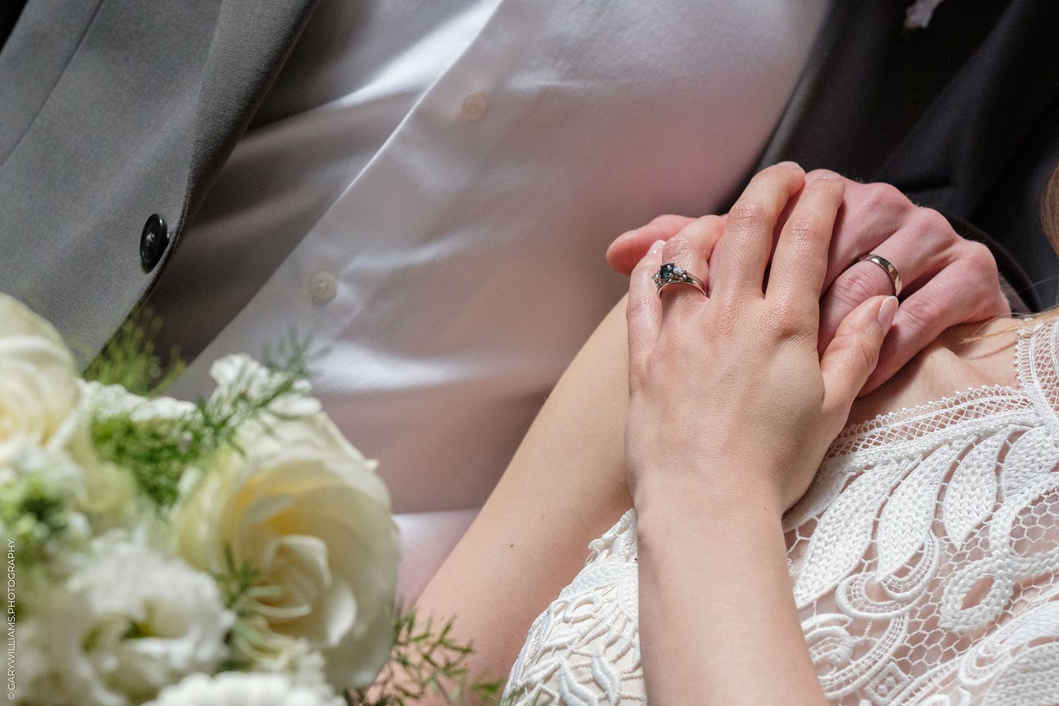 Hands clasped together on a white lace wedding dress beside cream roses and delicate floral arrangements.