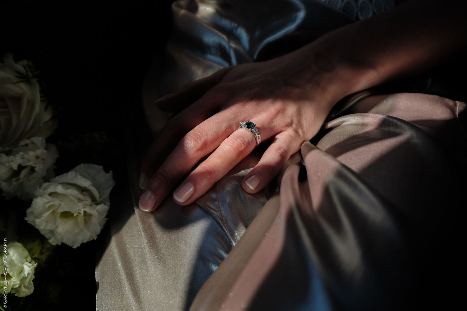 Close up of hands wearing engagement ring against formal attire with white roses in dark lighting.