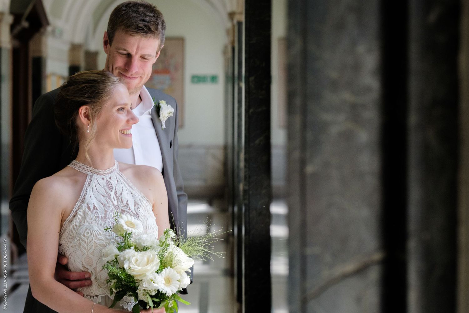 A bride and groom share an intimate moment in an ornate hallway as she holds a white floral bouquet against her lace dress.