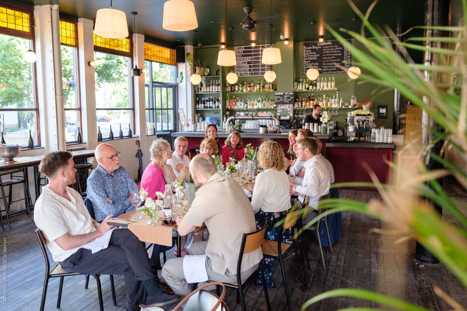 Restaurant patrons gather around a busy bar counter with pendant lights and large windows overlooking greenery outside.