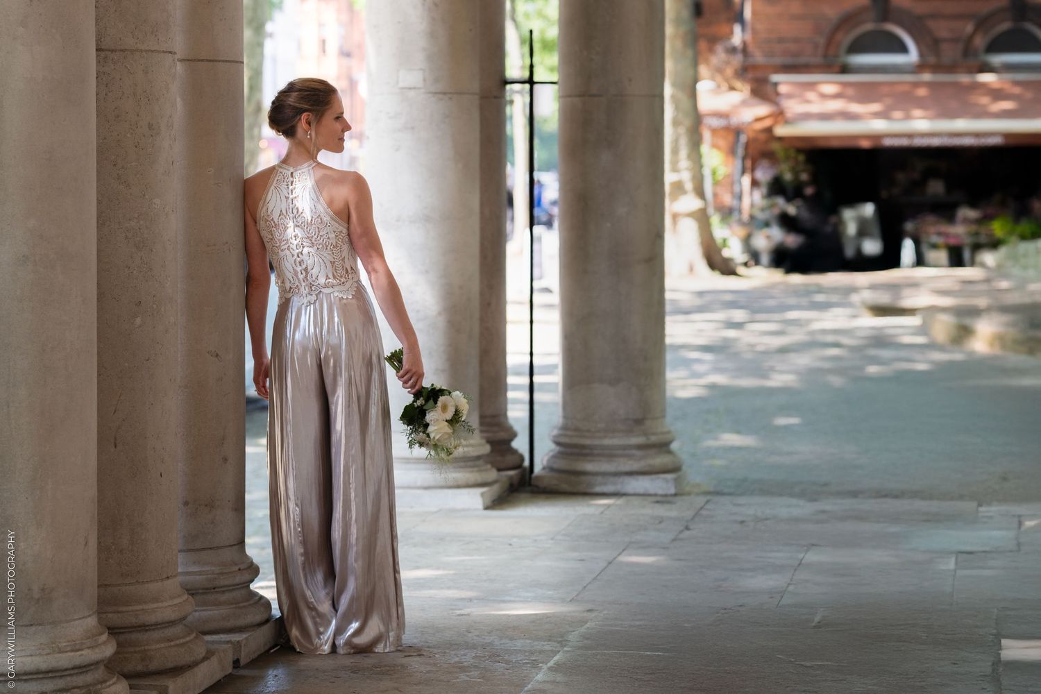 Figure in a silvery formal dress stands elegantly between stone columns holding a bouquet.