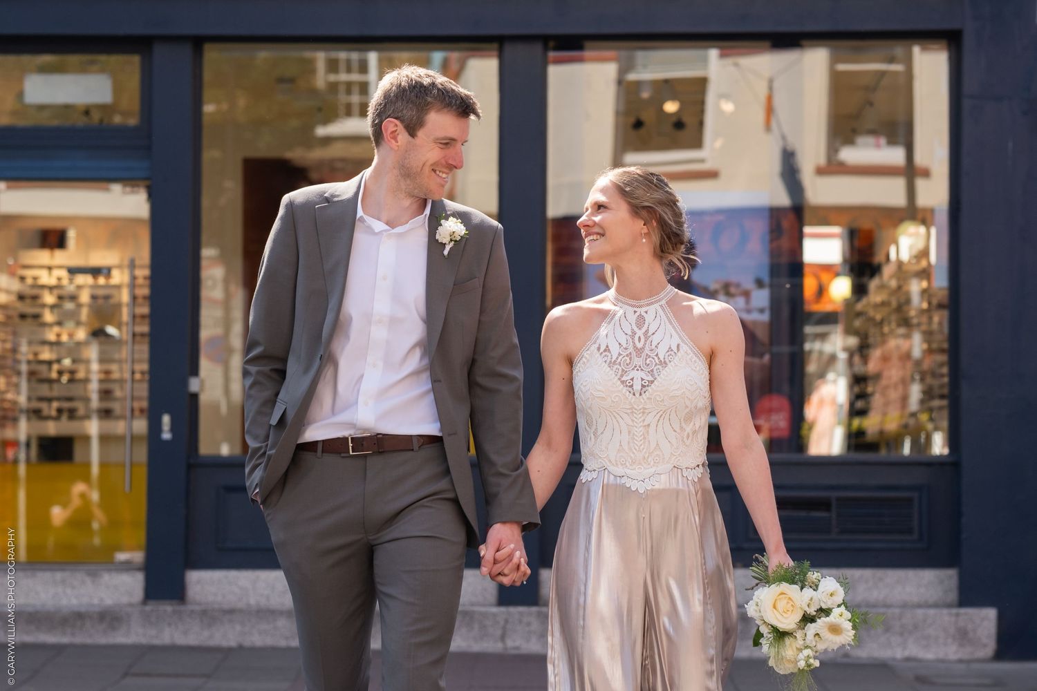 A bride and groom walk hand in hand on a city street with storefronts in the background.