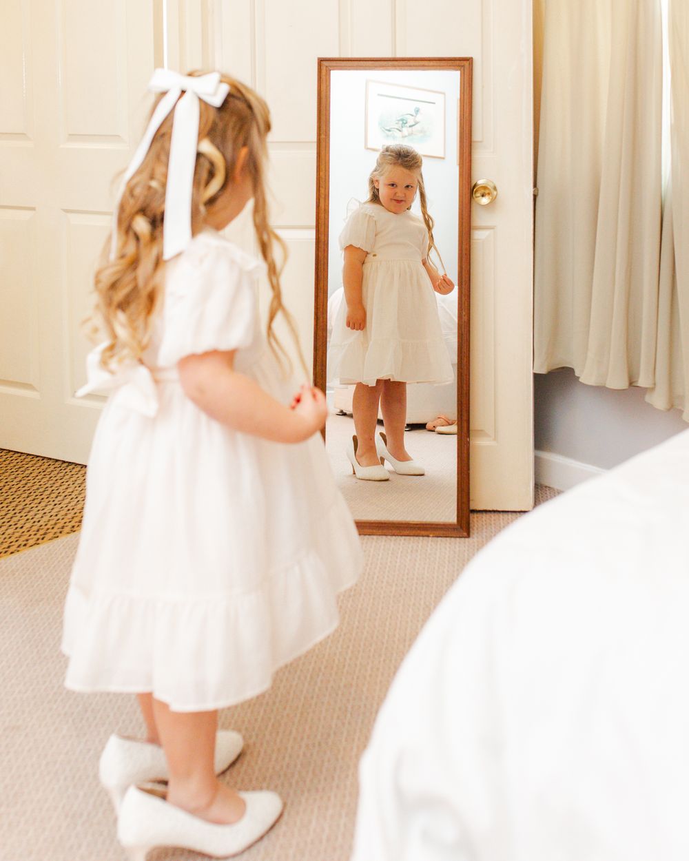 Young child in white dress with bow admiring reflection in full-length mirror on carpeted floor.