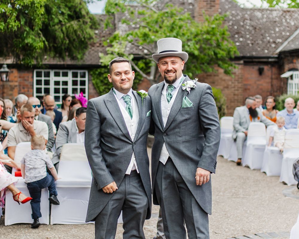 Two wedding attendees in formal morning suits with tails pose together at an outdoor reception with guests in background.