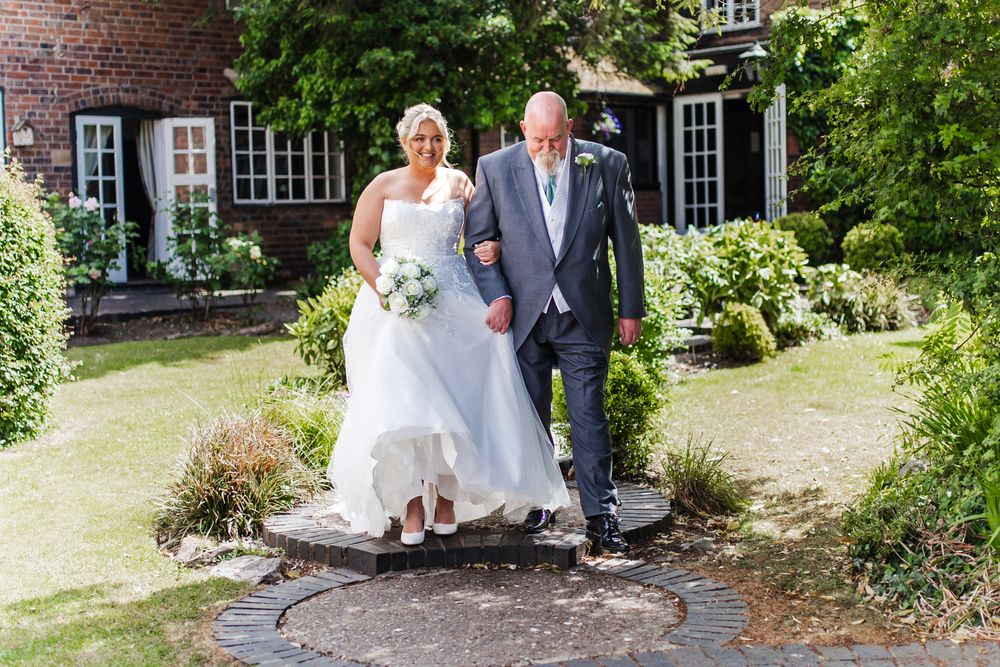 A couple in wedding attire walks down a garden path beside a charming brick house with white-trimmed windows.