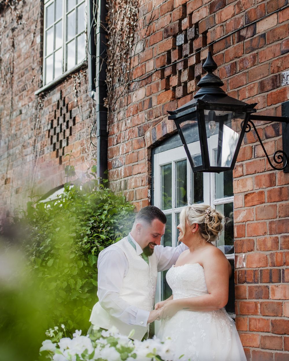 A romantic wedding couple poses against a rustic brick wall with decorative lantern and white florals at golden hour.
